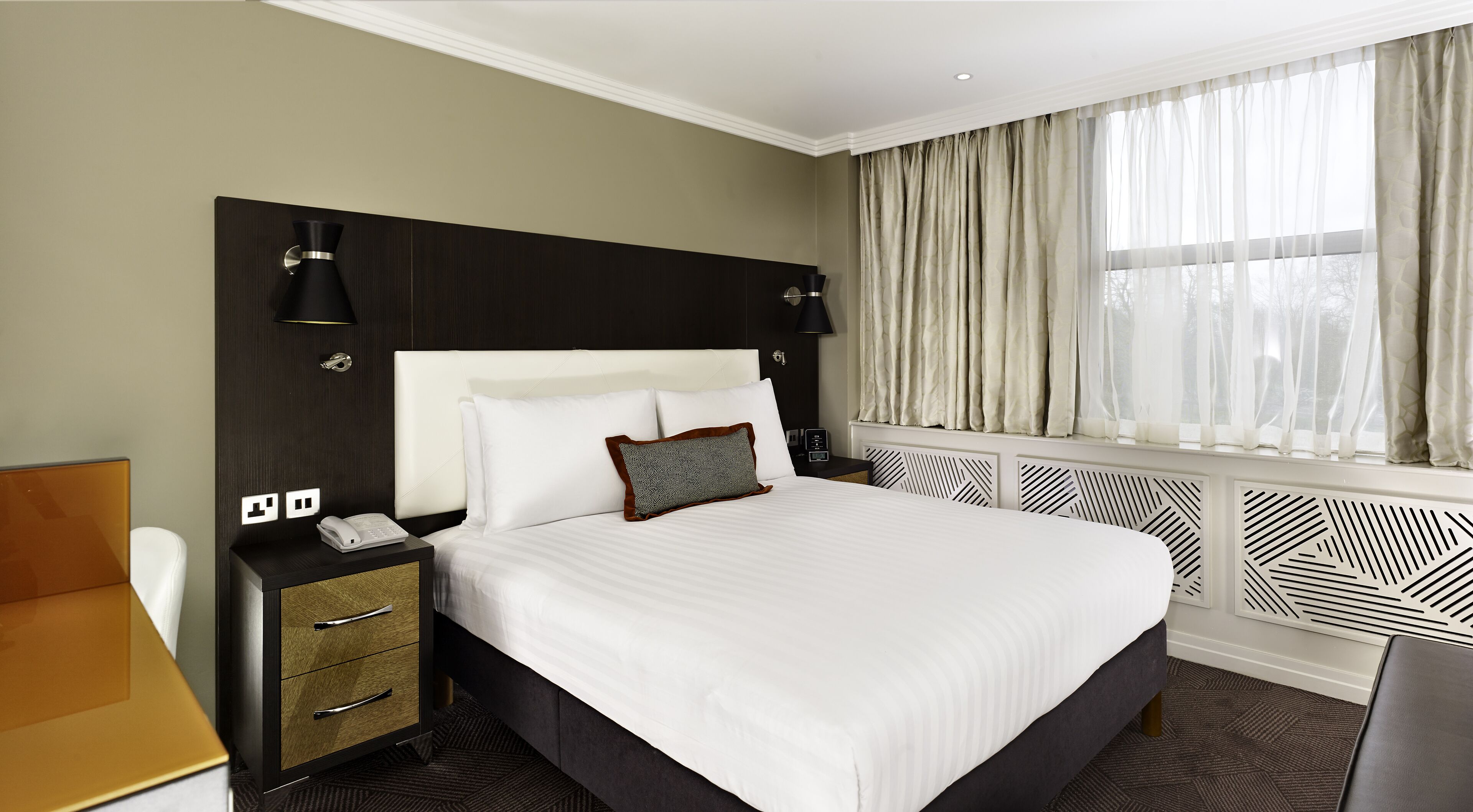 A hotel room with a large bed, bedside tables, a desk, and a window with curtains.