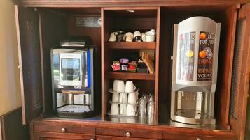 Una estació de cafè amb una màquina de cafè, un dispensador de te i una prestatgeria amb tasses i complements.