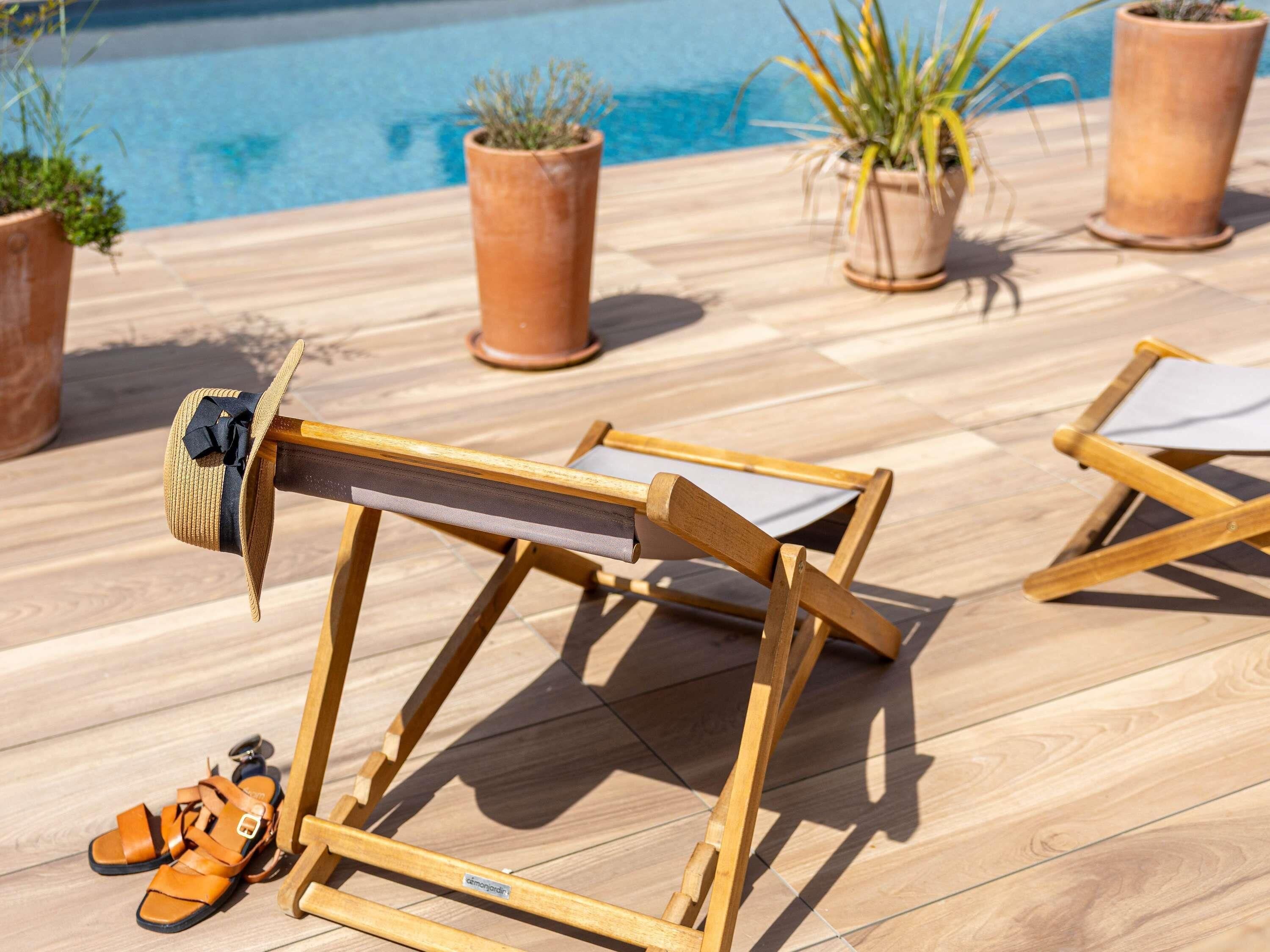 A wooden lounge chair with a sunshade, a pair of sandals, and potted plants near a pool.