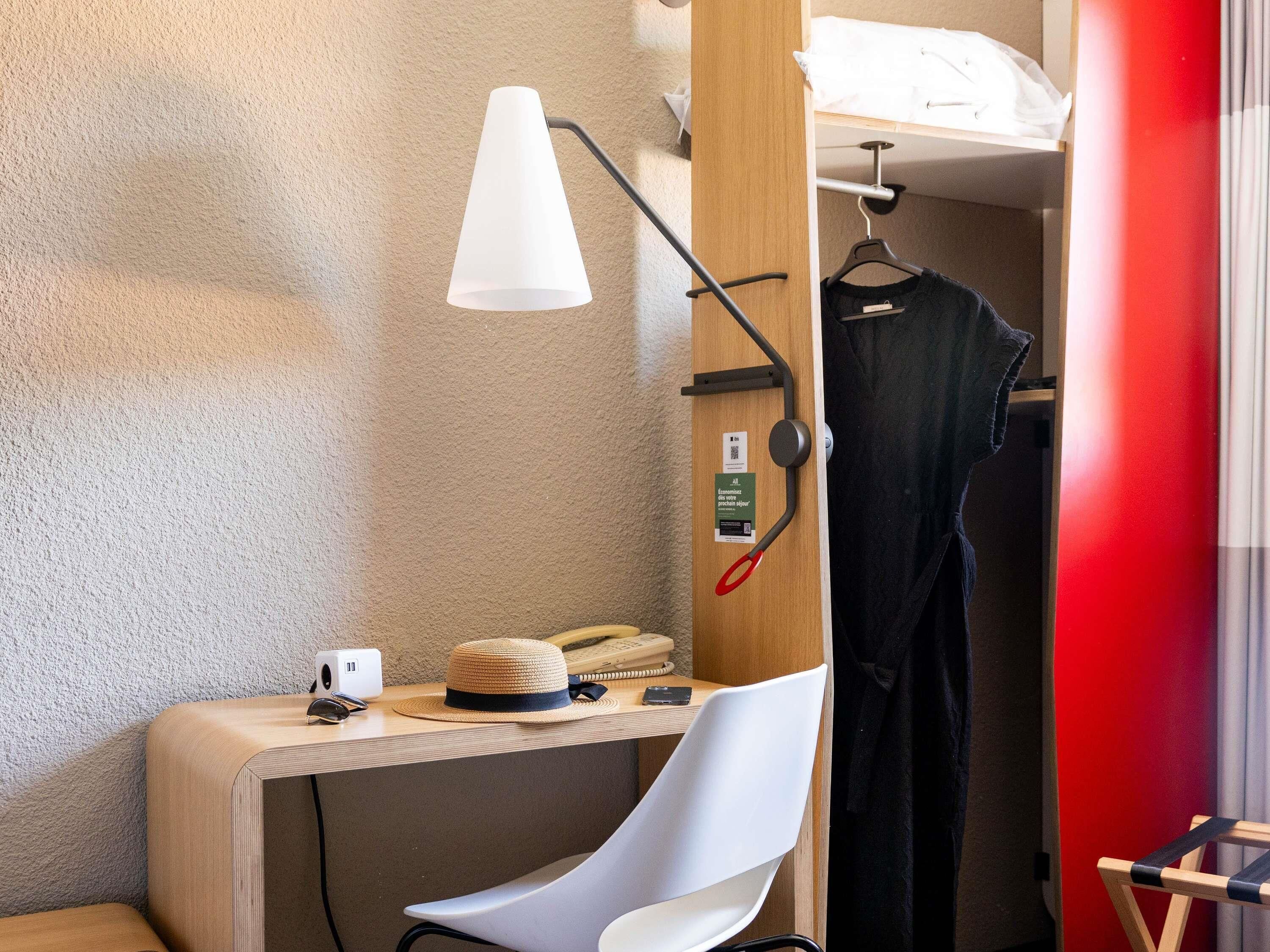 Habitación de hotel pequeña con escritorio, una silla blanca, perchero y lámpara de noche.
