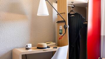 Habitación de hotel pequeña con escritorio, una silla blanca, perchero y lámpara de noche.