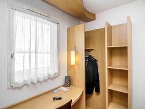 Une pièce avec un bureau en bois, une fenêtre ornée de rideaux légers, un porte-manteau avec une veste, et une armoire en bois dotée d’étagères.