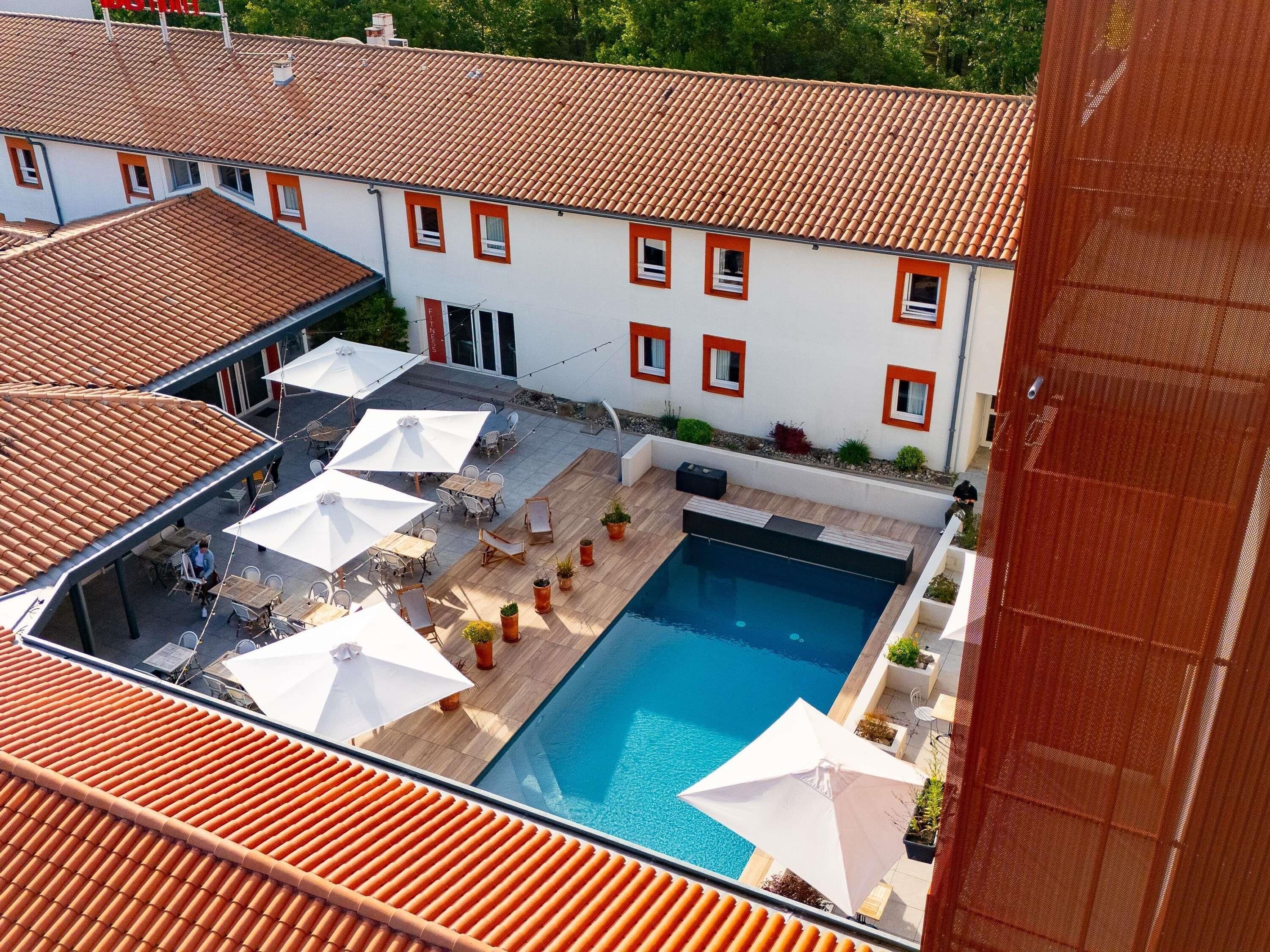 A rooftop pool area with outdoor seating and umbrellas, surrounded by buildings with red-tiled roofs.