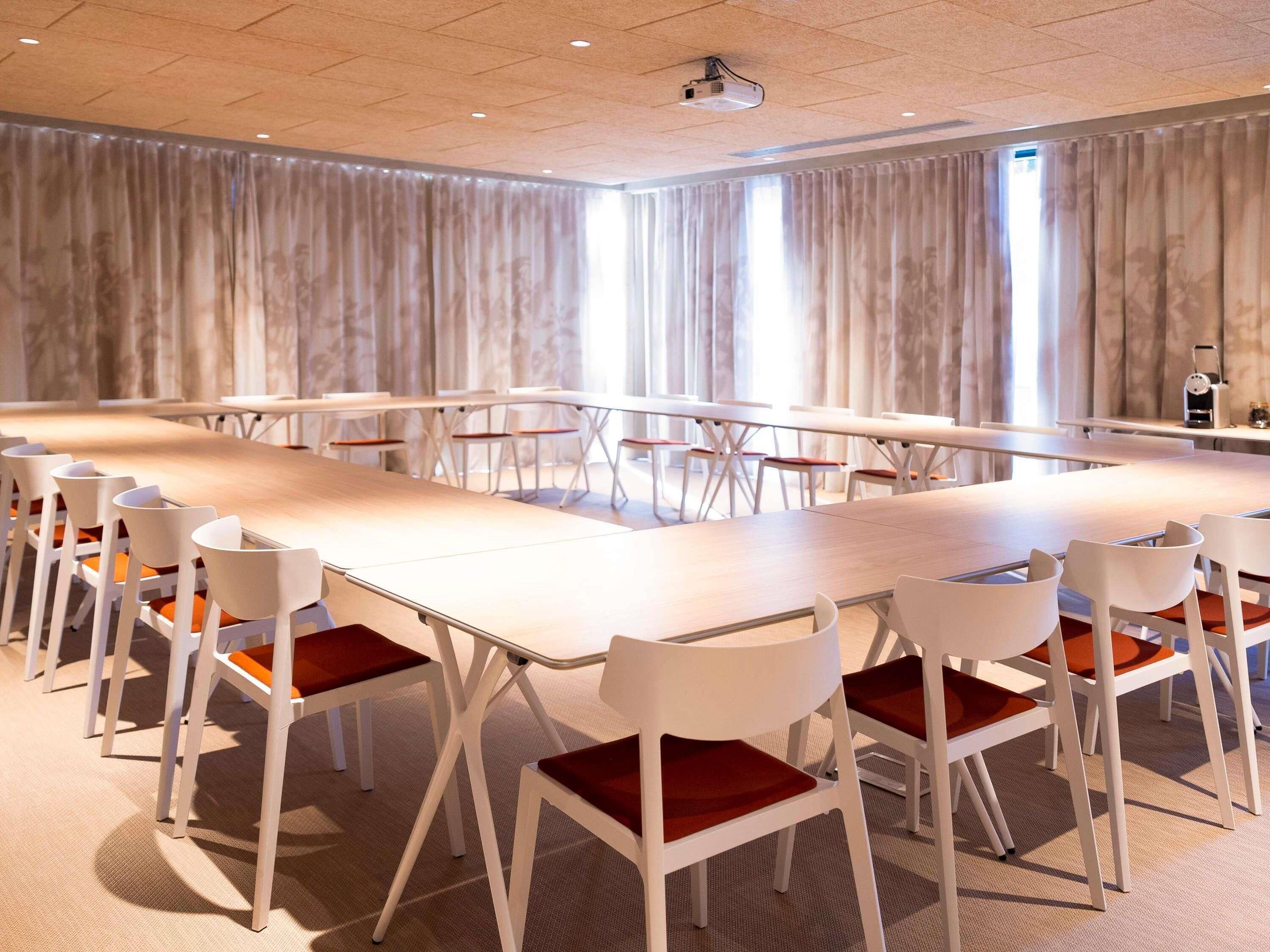 Un salón de conferencias con una mesa larga, sillas blancas con cojines rojos y un proyector en el techo.