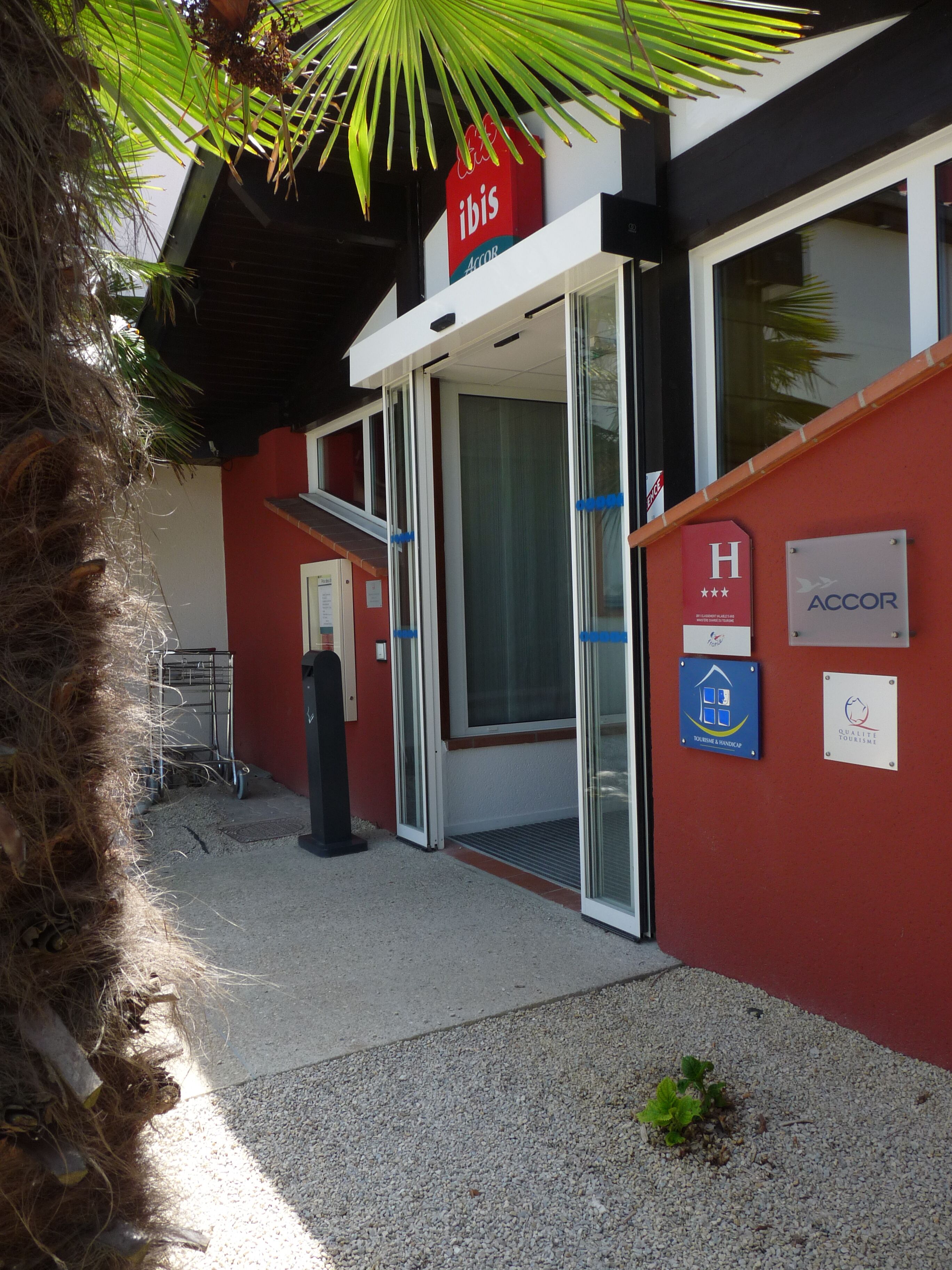Entrada d'hotel amb façana vermella, un senyal amb la marca 'ibis' i el símbol d'accessibilitat per a persones amb mobilitat reduïda.