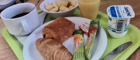 Mesa de café da manhã com croissants, uma tigela de frutas, um copo de suco e um pote de iogurte.