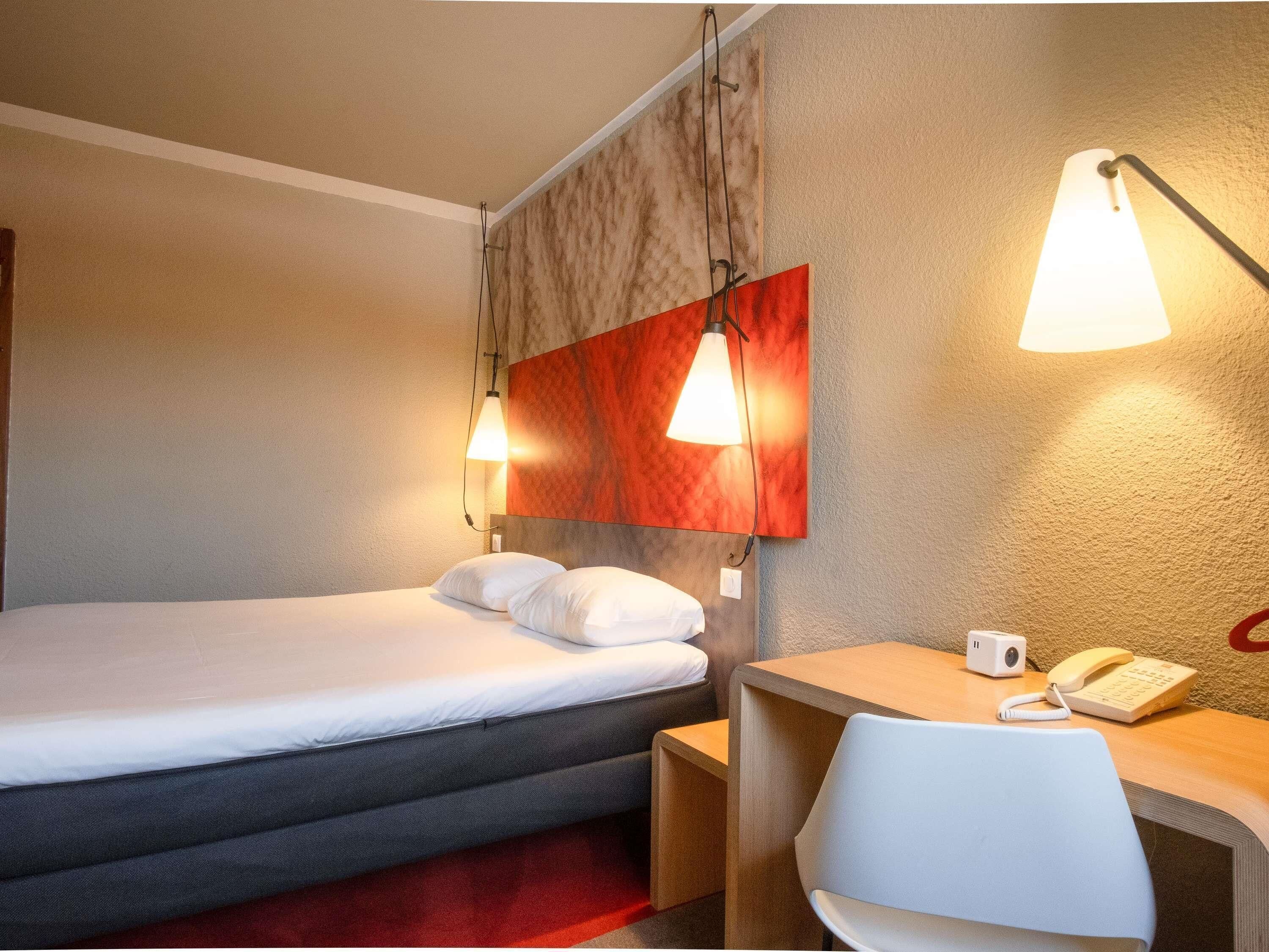 A hotel room with a bed, a desk with a phone, and two wall-mounted lamps.