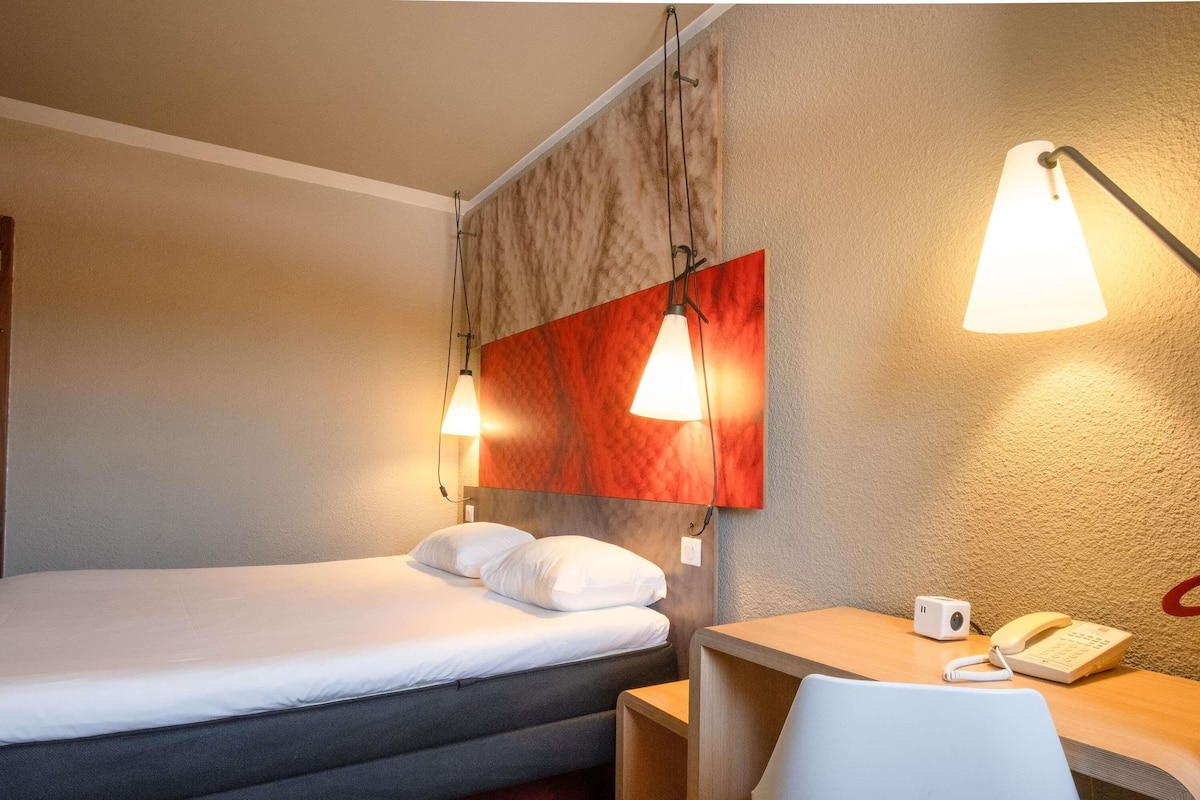 A hotel room with a bed, a desk with a phone, and two wall-mounted lamps.
