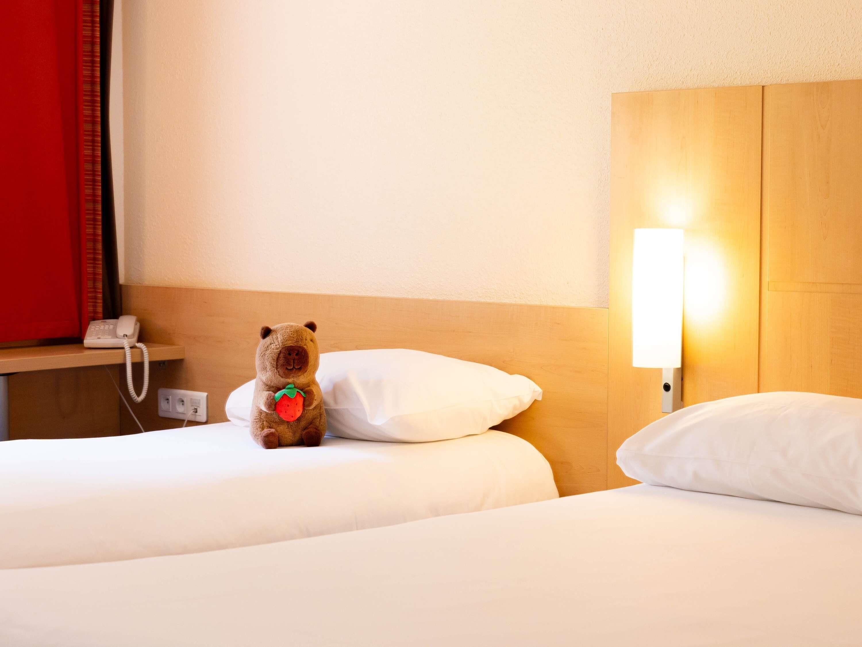 Une chambre d’hôtel avec deux lits, un téléphone sur la table de chevet, une lampe et un ours en peluche sur l’un des lits.