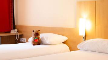 Une chambre d’hôtel avec deux lits, un téléphone sur la table de chevet, une lampe et un ours en peluche sur l’un des lits.