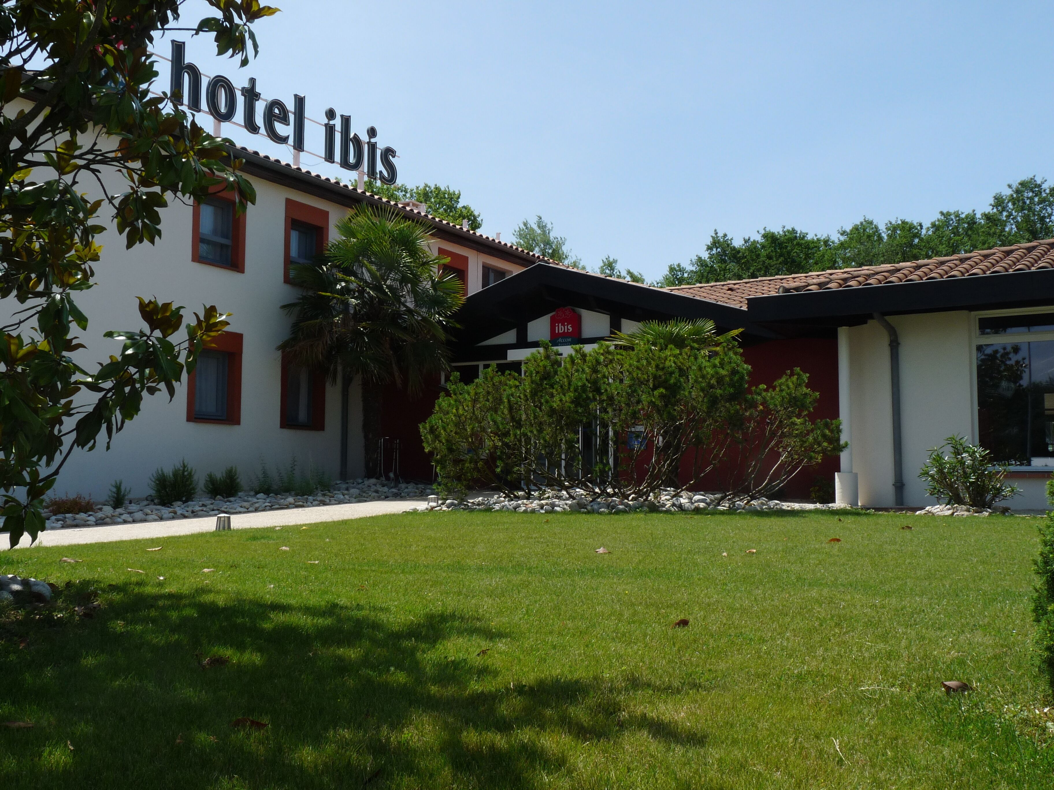 Ein Hotel namens 'hotel ibis' mit moderner Architektur, das sich durch ein flaches Dach, rote und weiße Außenwände sowie einen gepflegten Rasen mit kleinen Bäumen und Sträuchern auszeichnet.