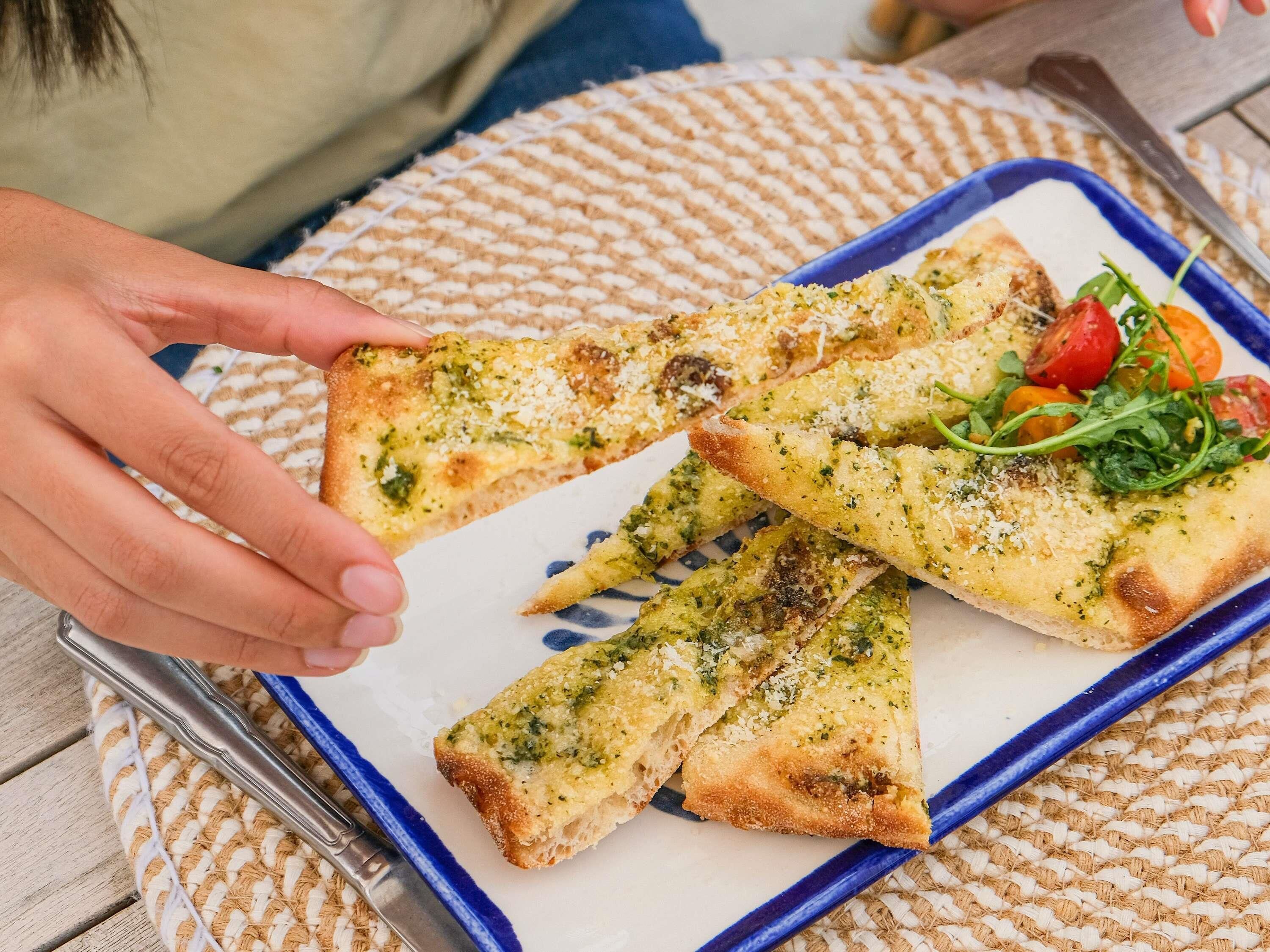 Une personne tenant une part de pizza recouverte d’une sauce pesto verte, servie dans une assiette bleue sur un fond blanc.