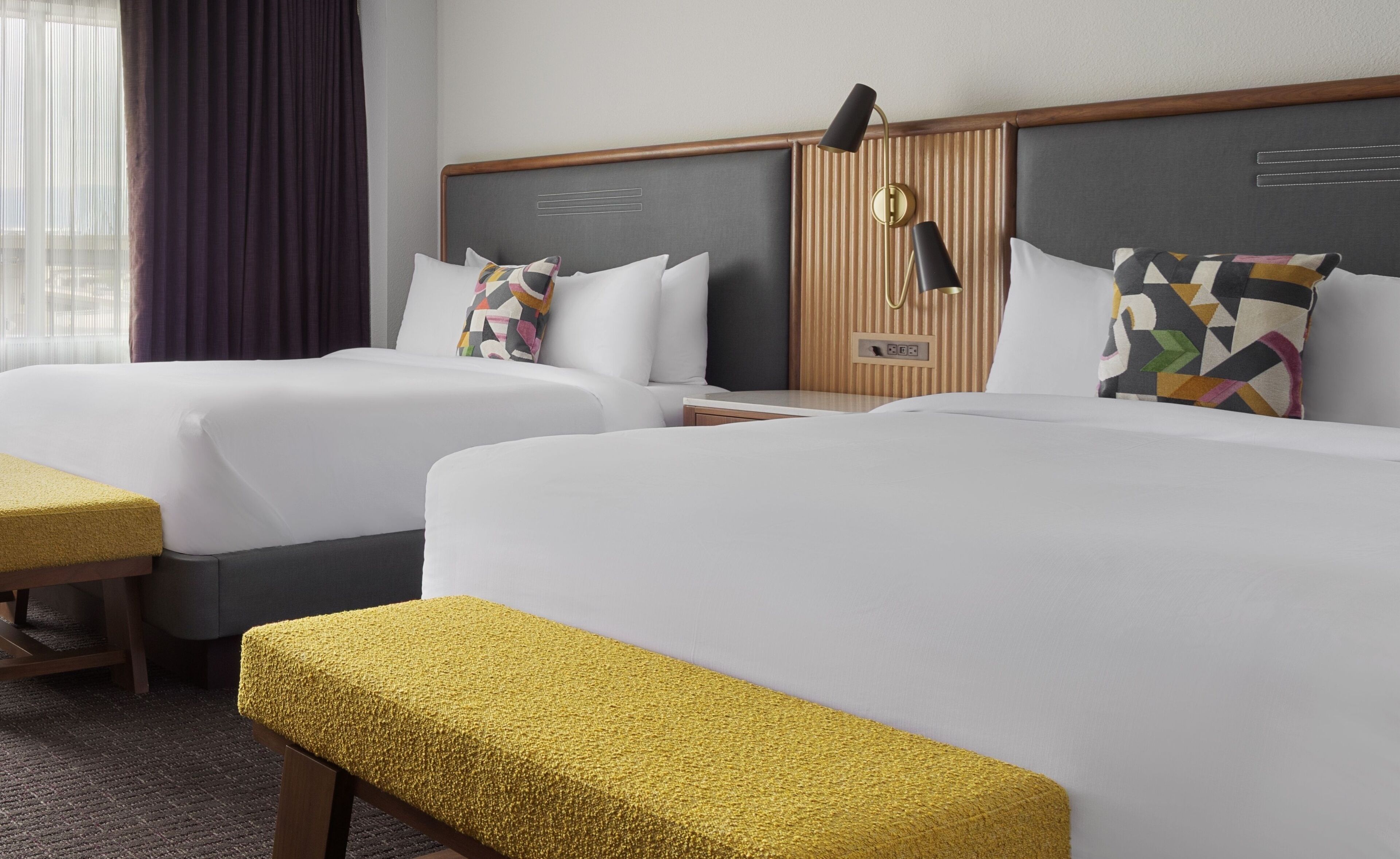 A hotel room with two beds, a bench, a bedside table, and a lamp.