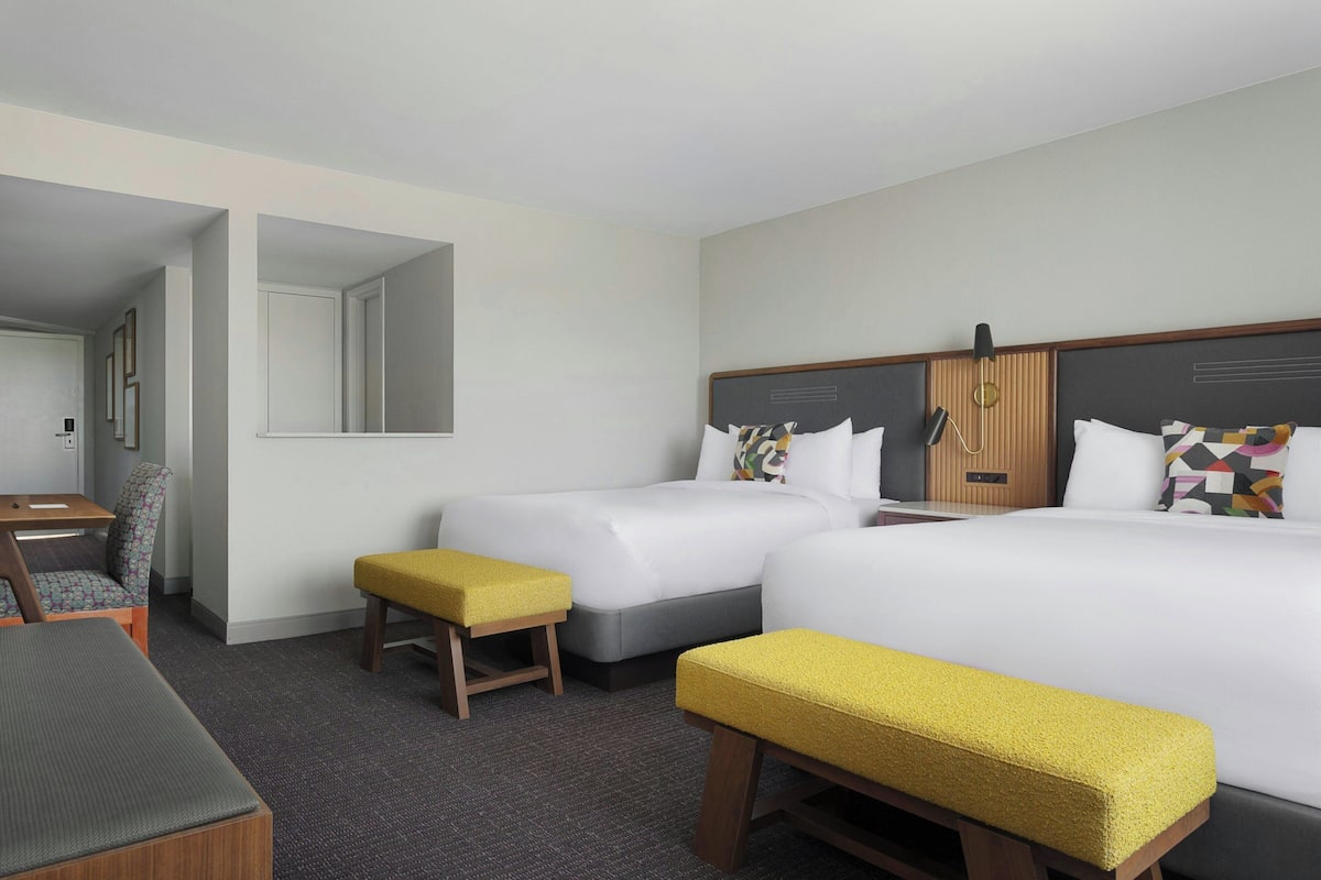 A hotel room with two beds, a desk, a chair, and a bench.
