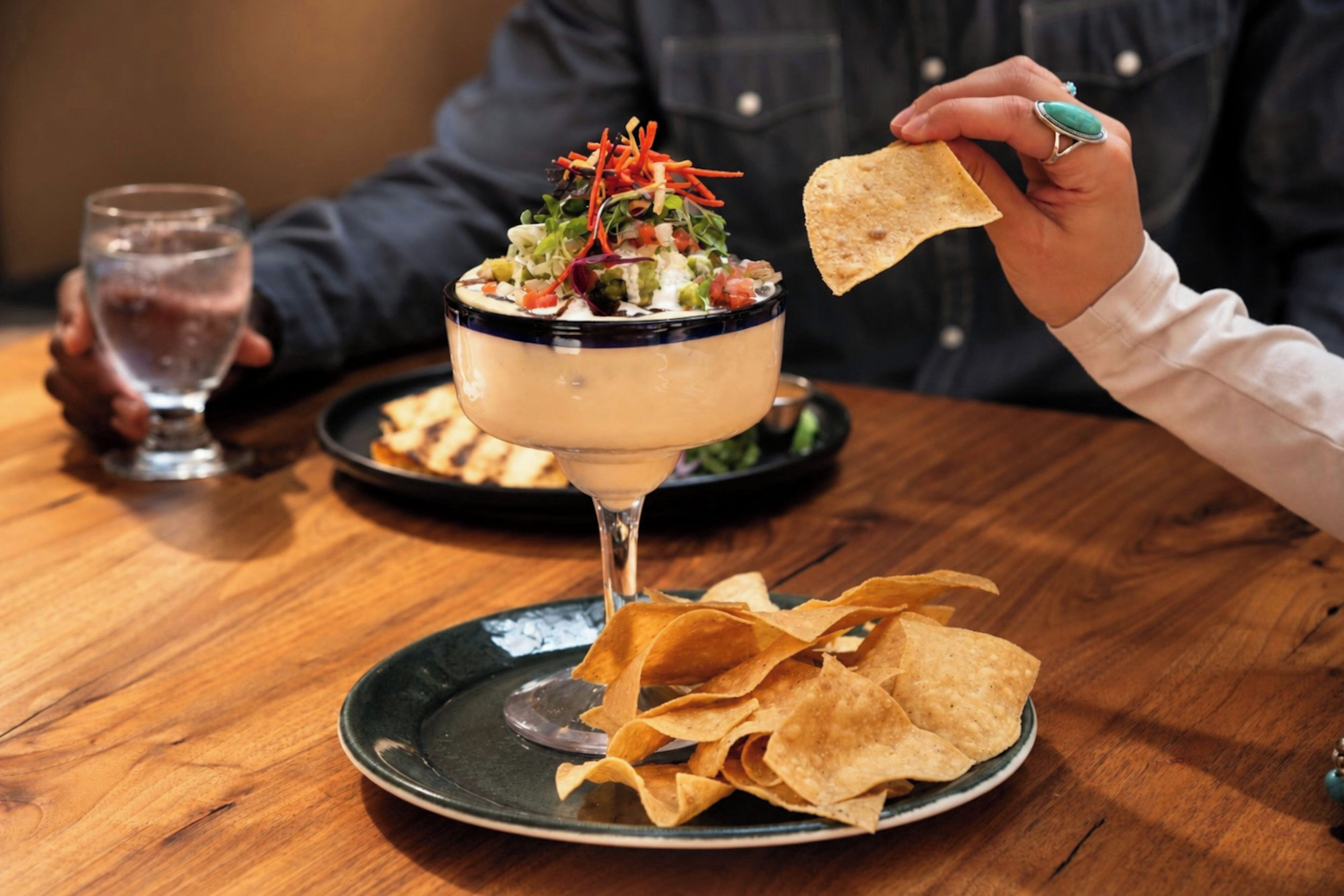 A person holding a tortilla chip over a bowl of salad with a drink in the background.