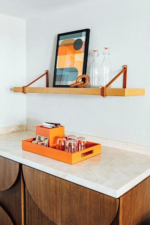 A kitchen counter with a wooden shelf holding a framed picture, two glass bottles, and a wicker basket. Below the shelf, there's an orange tray with glass jars and a card. - The Arlin, Arlington, a Tribute Portfolio Hotel (Arlington)
