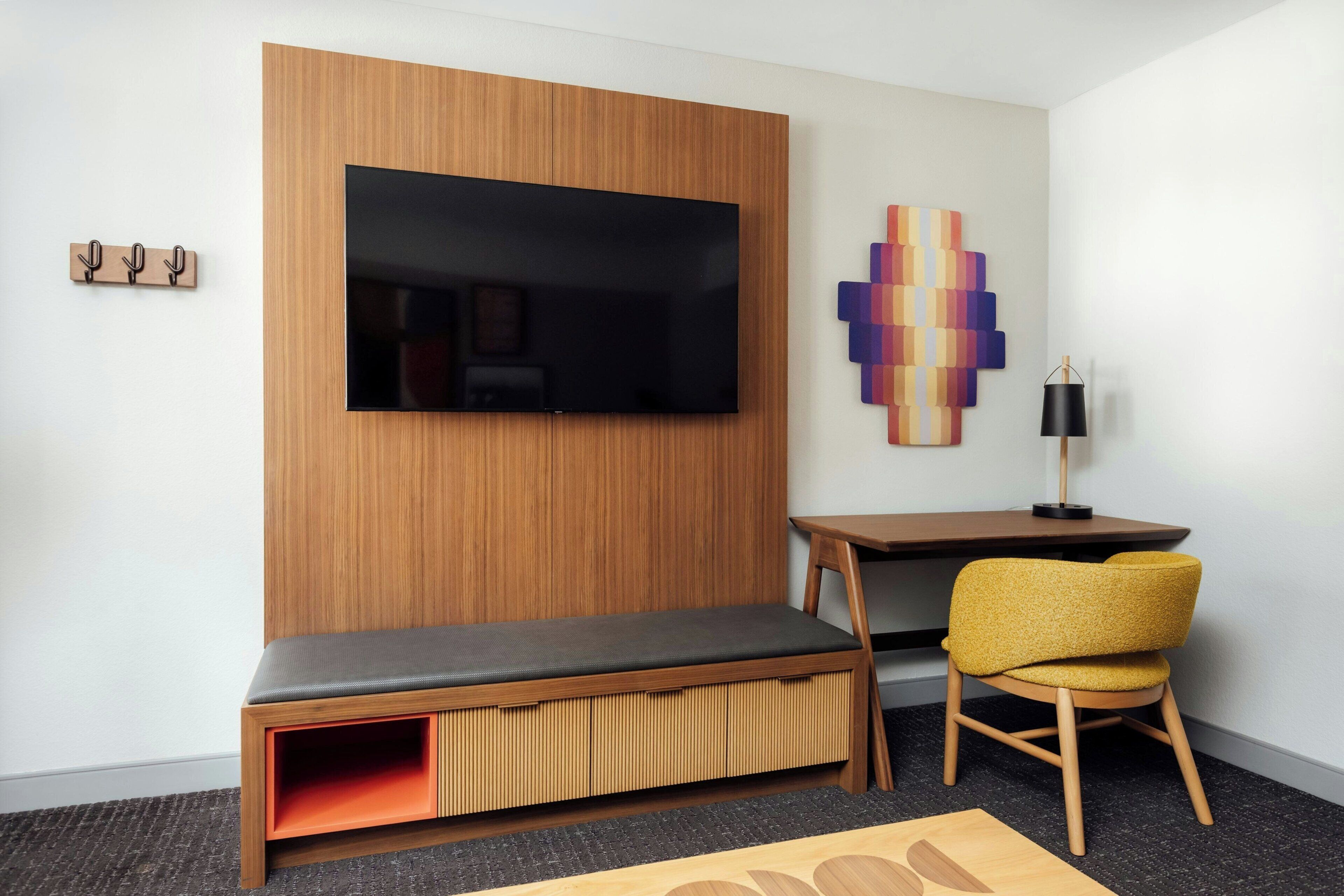 Ein modernes Hotelzimmer mit einer Holzwandverkleidung, einem Flachbildfernseher, einem farbenfrohen abstrakten Kunstwerk, einem kleinen Holztisch, einem gelben Sessel und einer Sitzbank mit Stauraum.