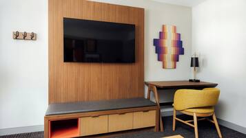 Ein modernes Hotelzimmer mit einer Holzwandverkleidung, einem Flachbildfernseher, einem farbenfrohen abstrakten Kunstwerk, einem kleinen Holztisch, einem gelben Sessel und einer Sitzbank mit Stauraum.