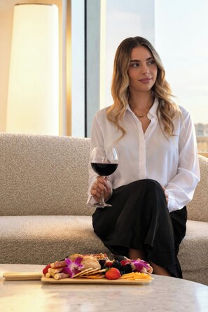 Una persona sentada en un sofá con una copa de vino tinto, un plato de comida y una tabla de madera con varios objetos.