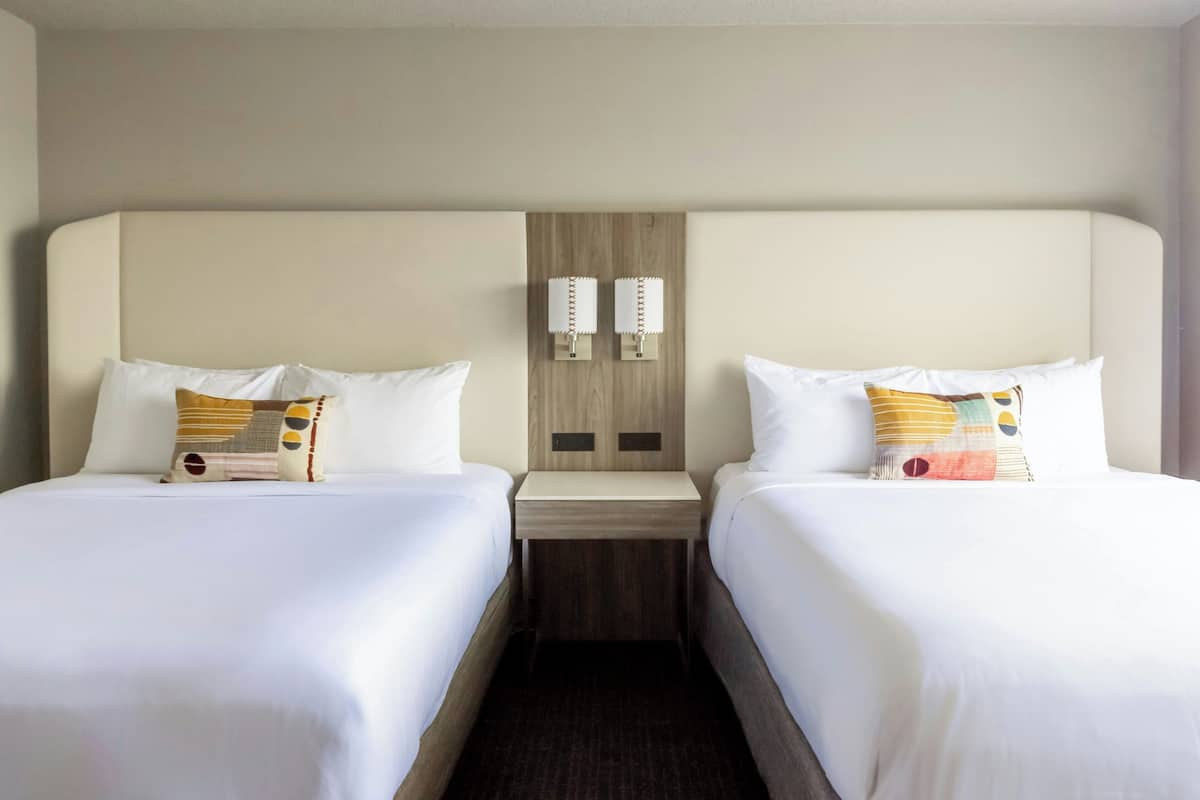 A hotel room with two beds, a wooden headboard, a nightstand, and two wall-mounted lamps.