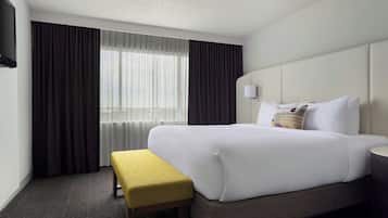 Una habitación de hotel con una cama grande, una mesita de noche, un banco, un televisor y una ventana con cortinas.