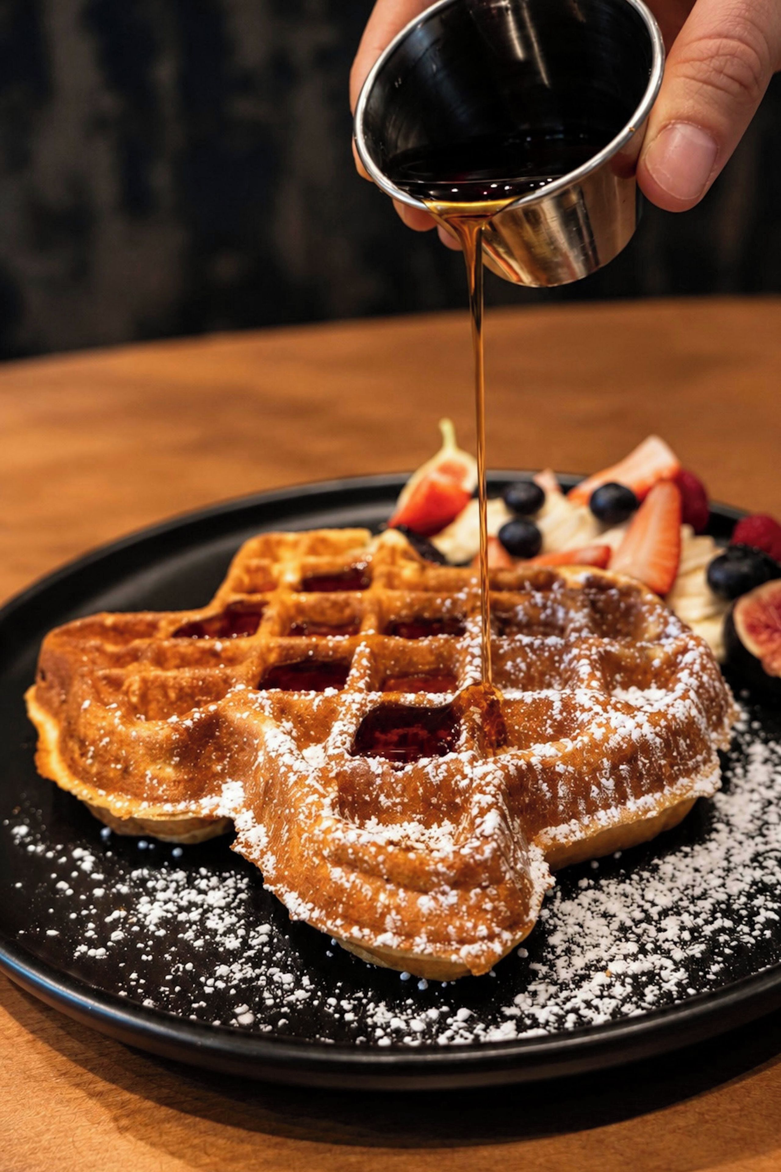 Un piatto con waffle ricoperti di zucchero a velo e sciroppo, accompagnati da frutti di bosco freschi e una fetta di torta.