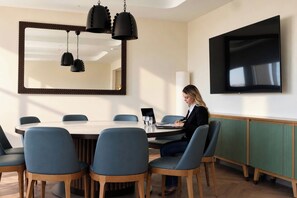 A conference room with a large table, blue chairs, a person seated, a large mirror, and a flat-screen TV on the wall.