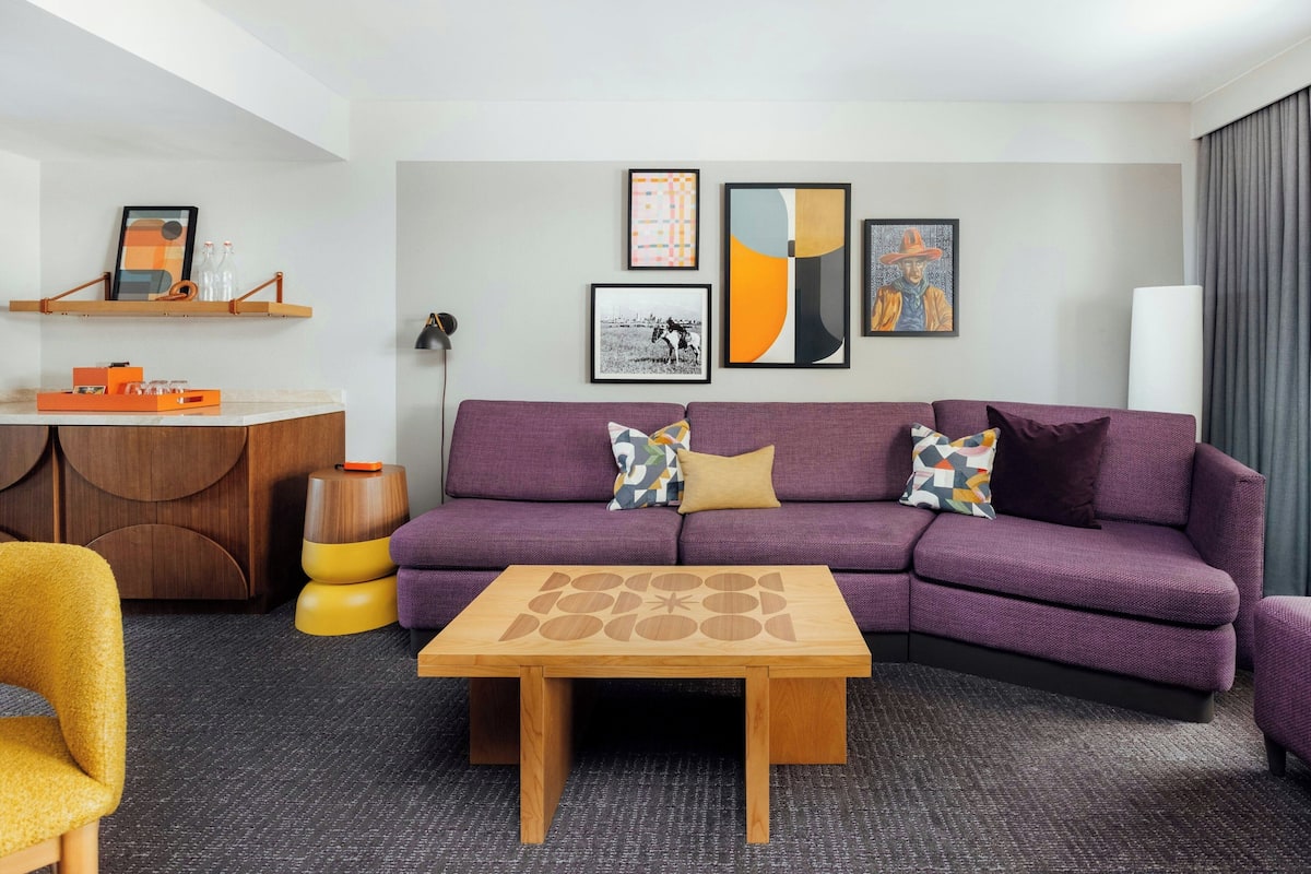 A modern living room with a purple sofa, a wooden coffee table, and a small side table. There are framed artworks on the wall and a desk with a chair.