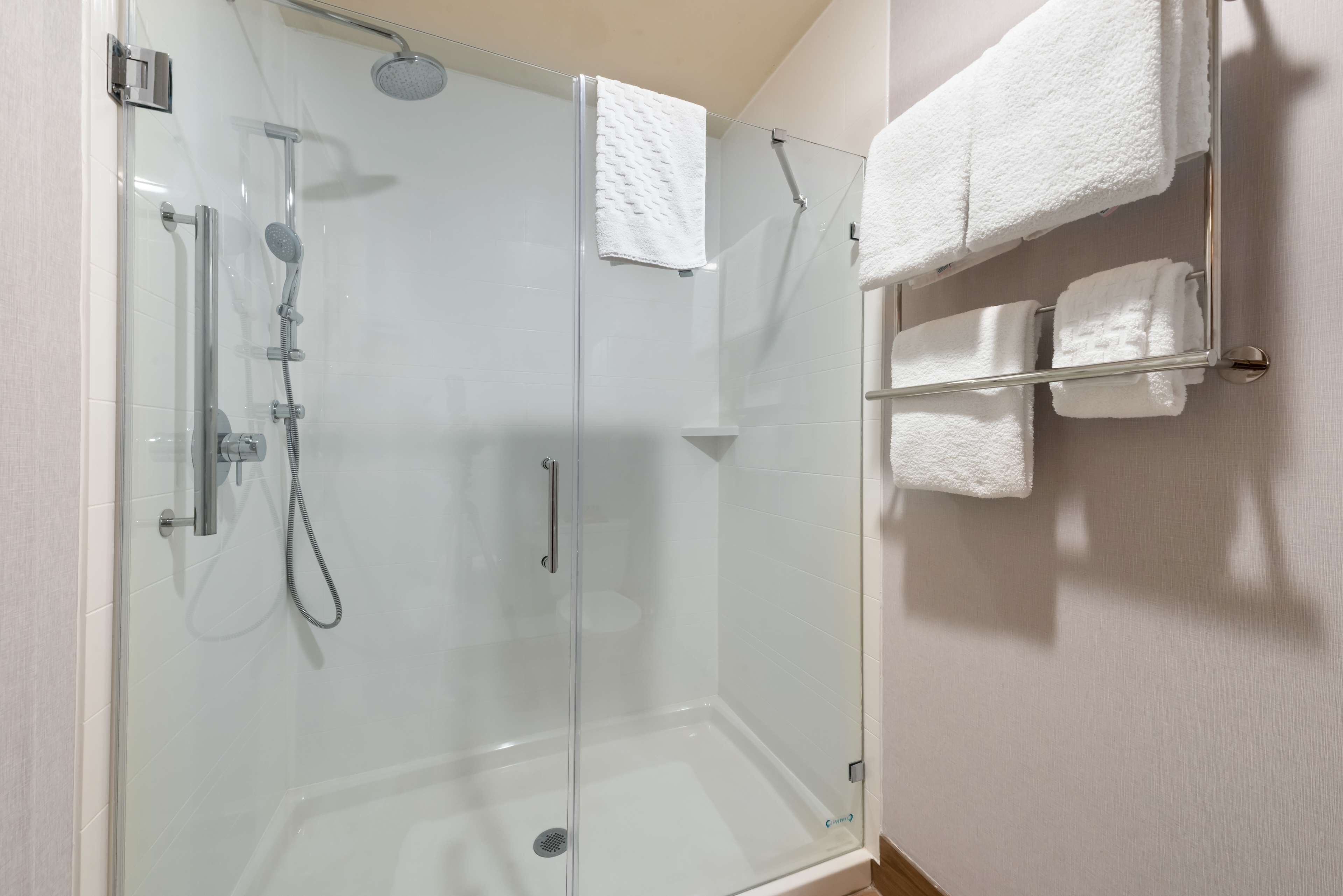 A glass-enclosed shower with a handheld showerhead, a handheld shower nozzle, and a wall-mounted towel rack with several white towels.