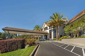 A hotel entrance with a covered walkway, parking lot, and palm trees.