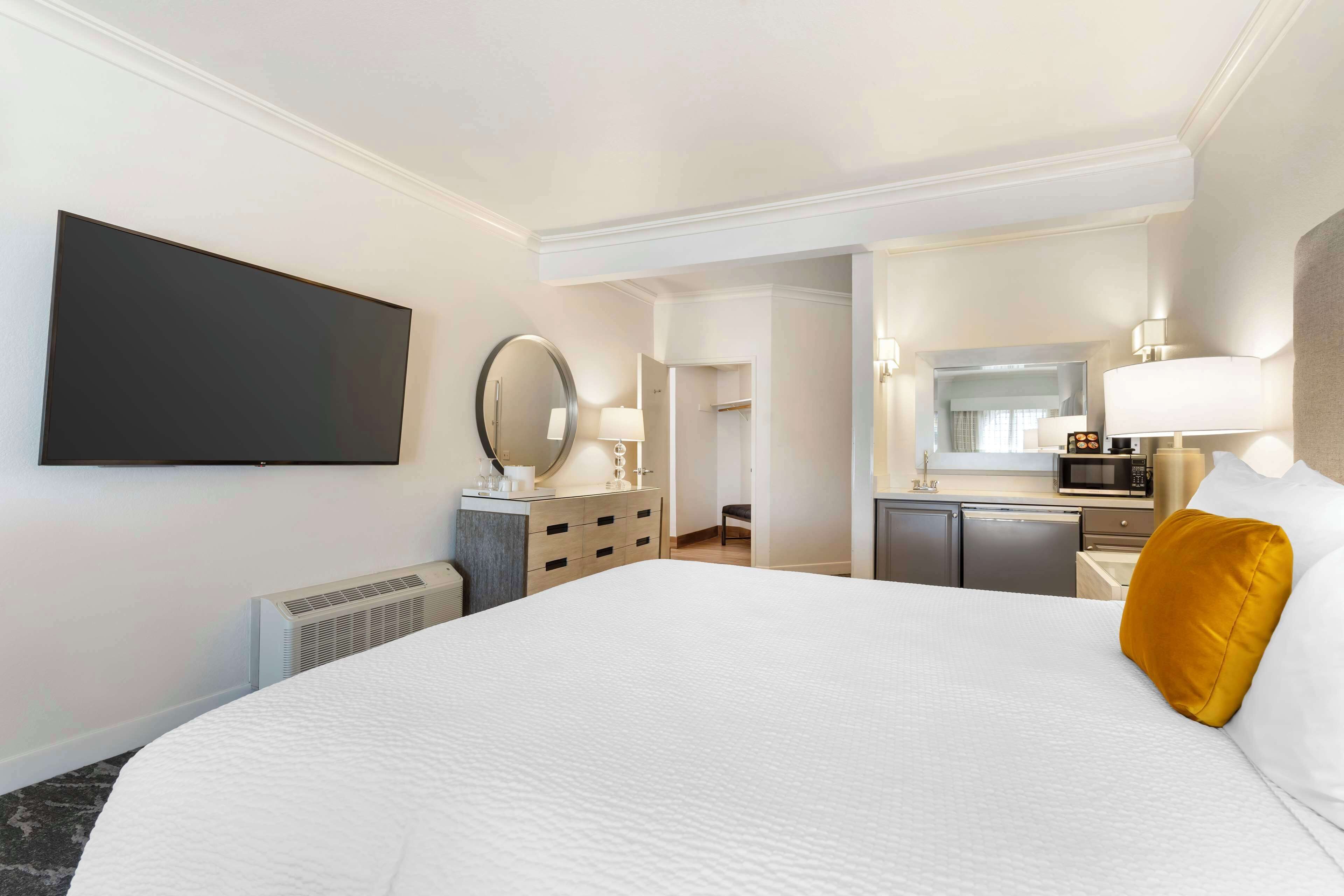 Habitación de hotel con una cama grande, un televisor de pantalla plana en la pared, una mesita de noche con lámpara y vista a la habitación contigua.
