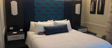 Cama bem arrumada com roupa de cama branca e um travesseiro azul de destaque, com duas mesas de cabeceira de cada lado, com luminárias, um relógio e um telefone.