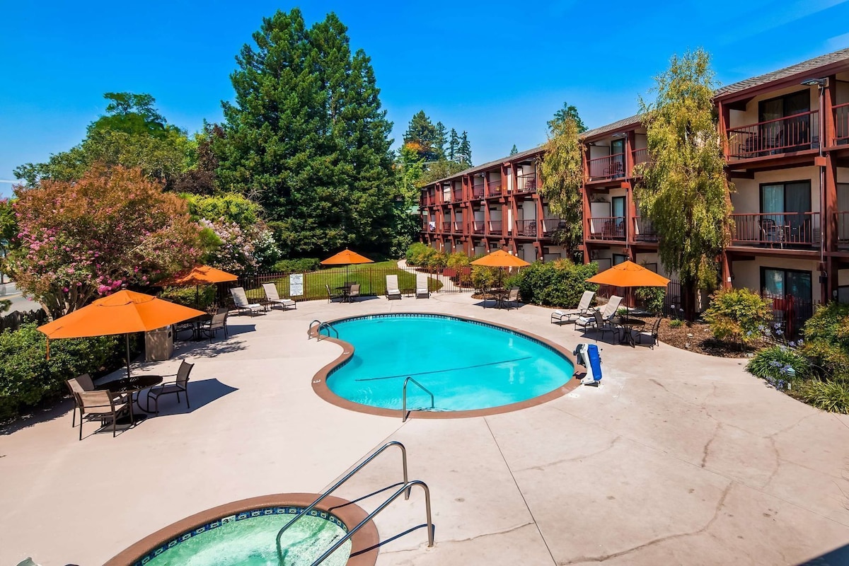 A swimming pool with lounge chairs and umbrellas, surrounded by trees and a building with balconies.