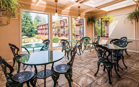 A sunlit patio with glass tables and chairs, large windows offering views of a building and trees.