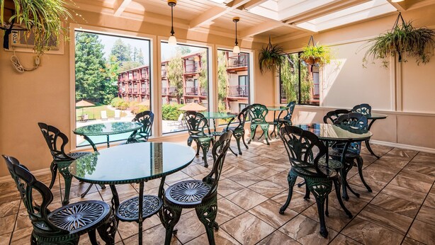 Une terrasse ensoleillée, dotée de tables et de chaises en verre, et de grandes fenêtres offrant une vue sur un immeuble et des arbres.