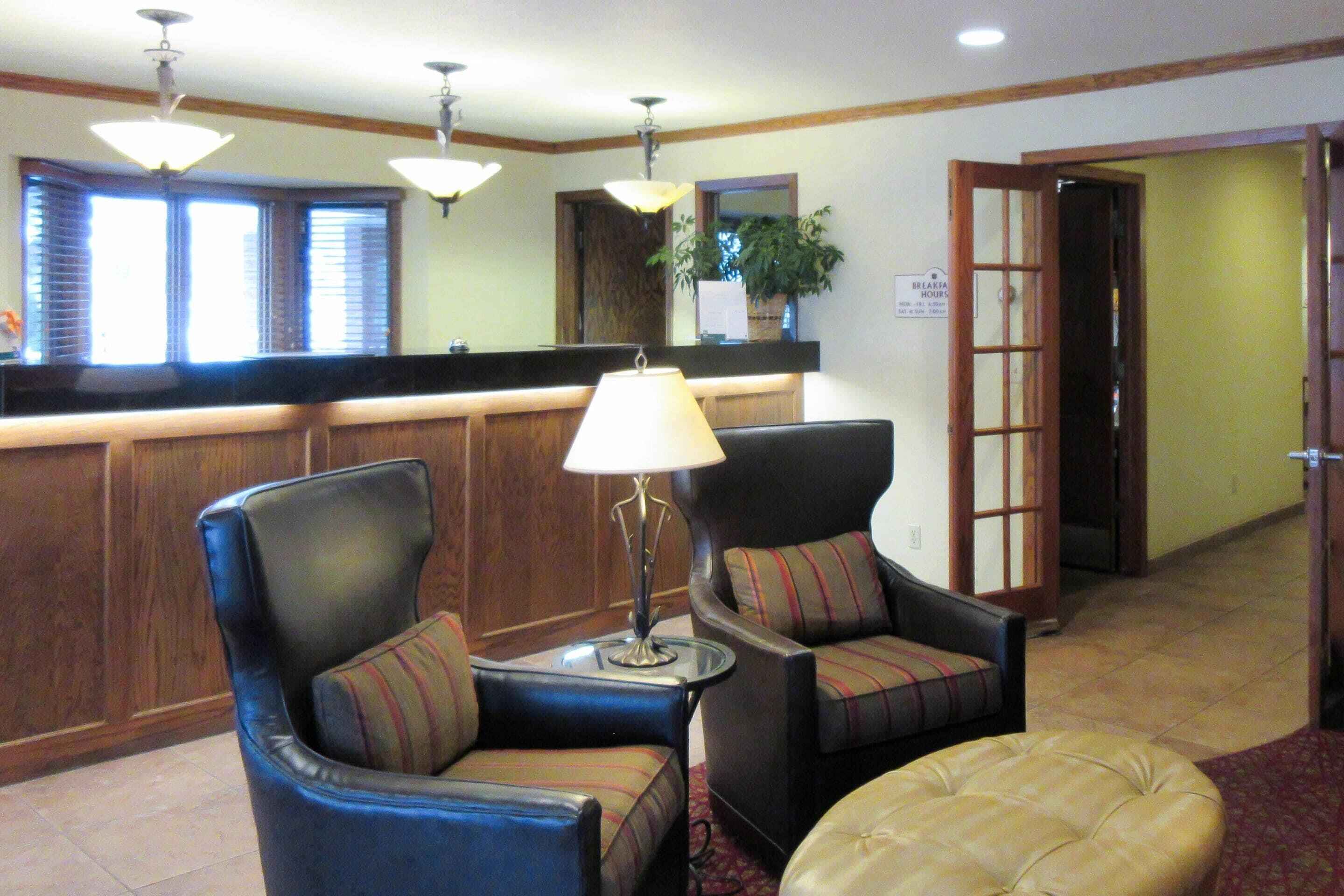 A lobby with two leather armchairs, a small table with a lamp, a reception desk, and a sign indicating breakfast hours.