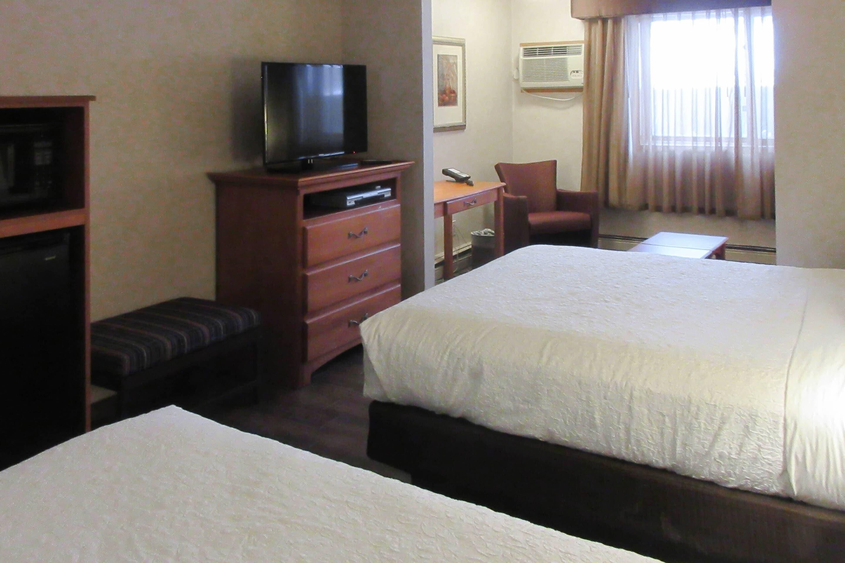 A hotel room with two beds, a TV on a wooden dresser, a desk with a chair, and a window with curtains.