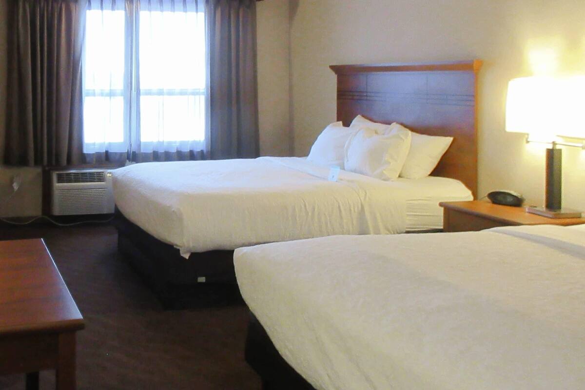 A hotel room with two beds, a desk, a lamp, and an air conditioning unit.