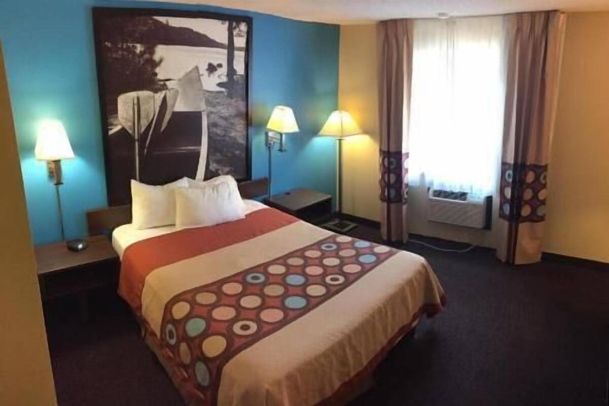 A hotel room with a bed, two bedside lamps, a nightstand, a large abstract painting on the wall, and a window with curtains.
