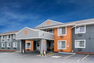 Un hotel de varios pisos con fachada gris y naranja, un acceso cubierto y un estacionamiento con espacios delimitados.