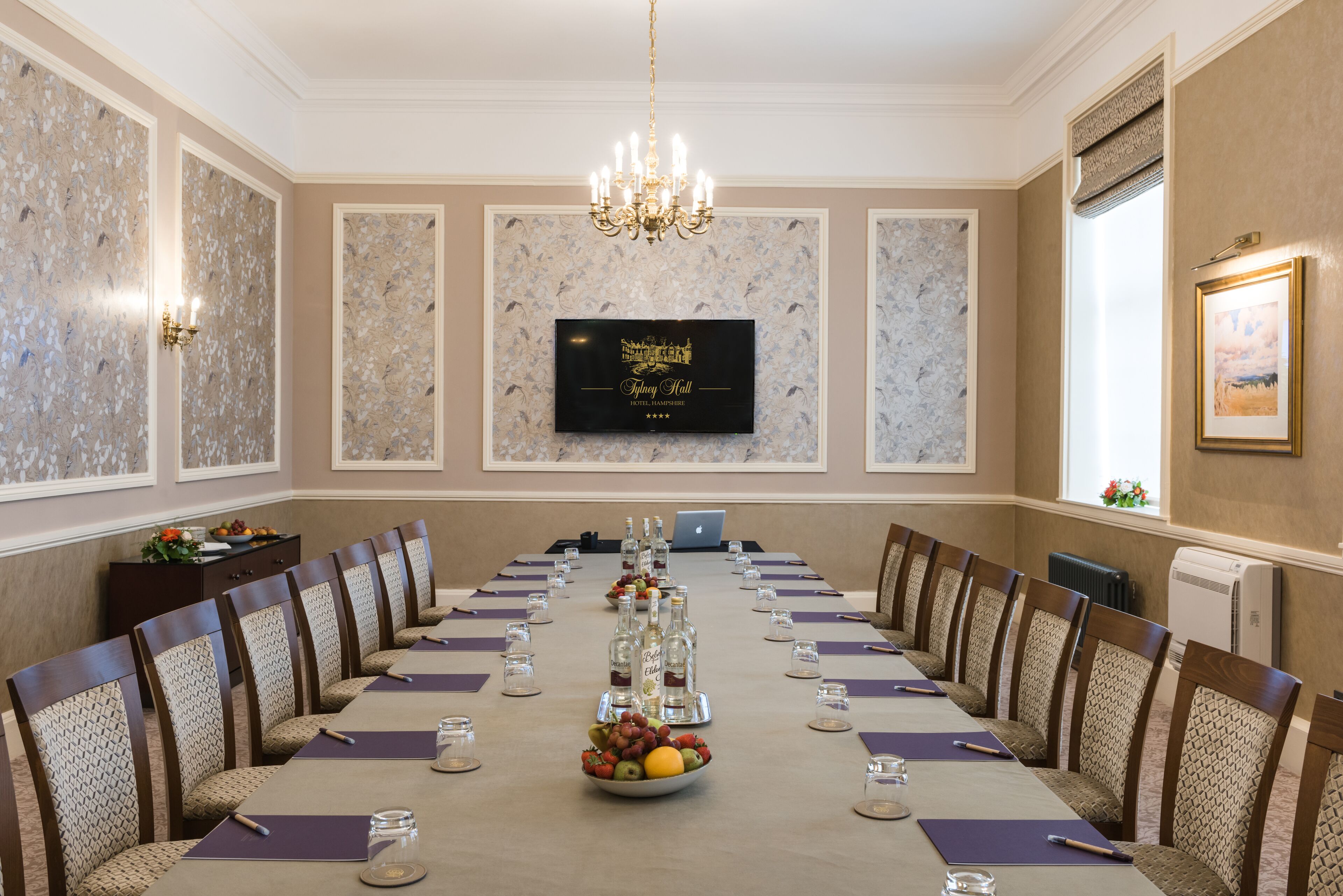 A conference room with a long table, chairs, a chandelier, a television screen, and a bowl of fruit.