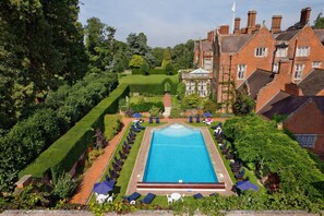 A large rectangular pool surrounded by lounge chairs and umbrellas, with a well-manicured lawn and a brick building in the background.