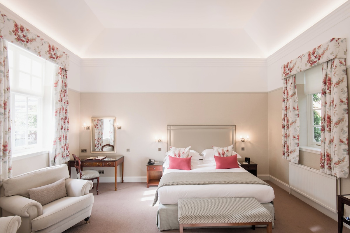 A hotel room with a bed, a desk, a chair, a mirror, and a window with floral curtains.