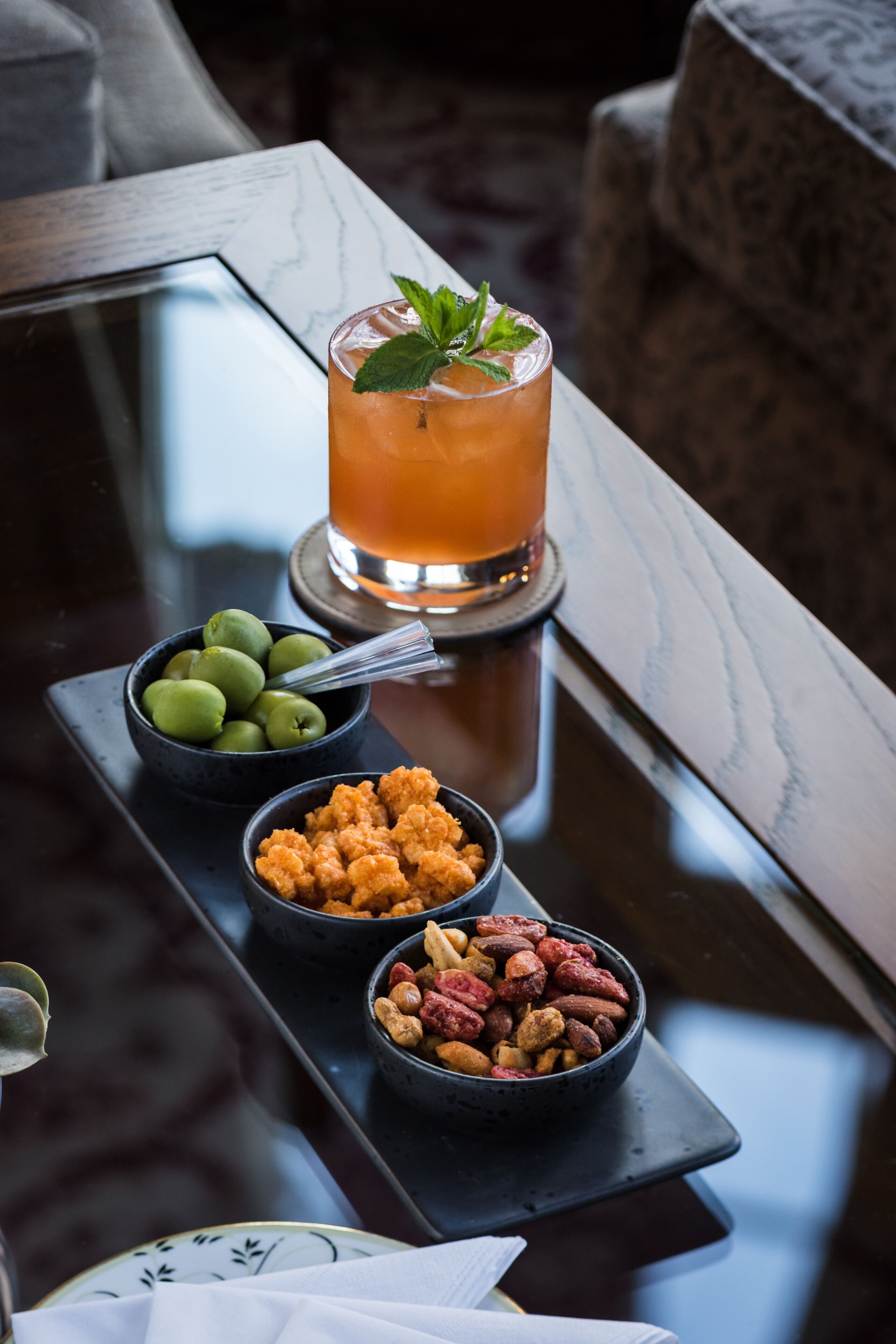 A tray with a drink, olives, and nuts on a glass table.