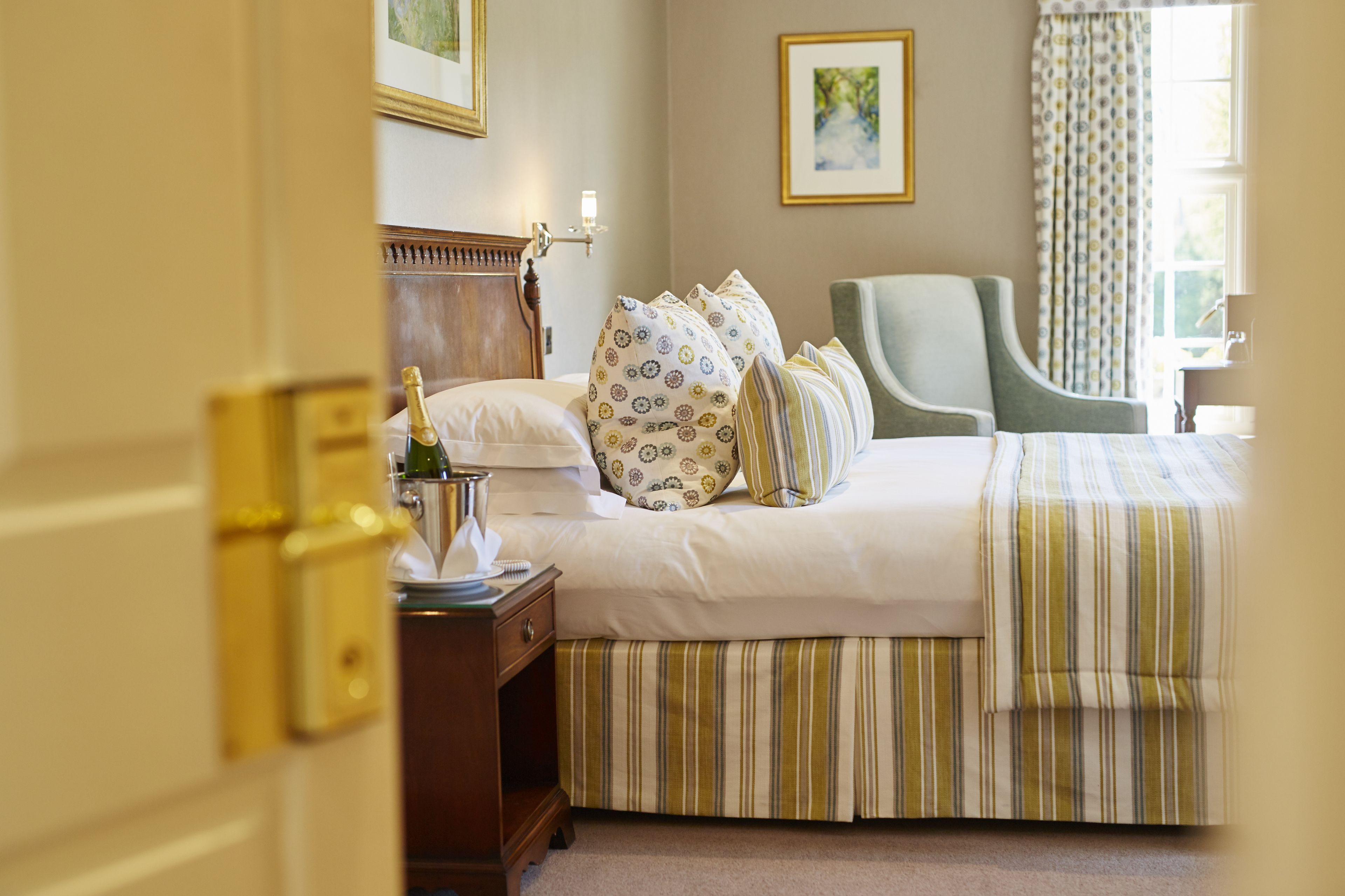 A neatly made bed with a striped bedspread, a bedside table with a bottle and a tissue box, and a chair in a well-lit room with a window and curtains.