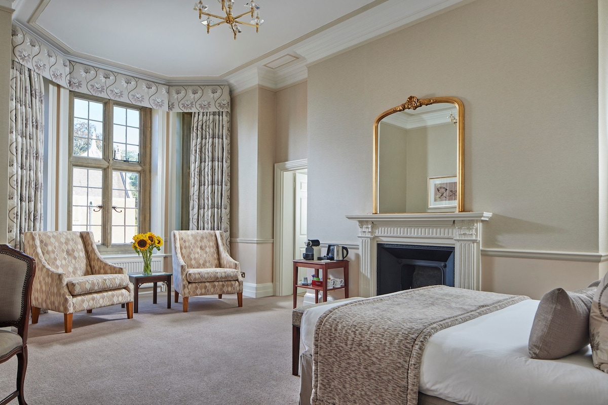 A hotel room with a large bed, two armchairs, a small table with a vase of sunflowers, a fireplace, and a large mirror.