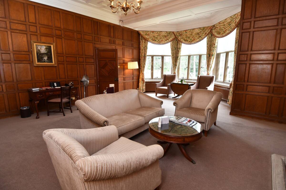A room with a wooden paneled wall, a desk with a chair, two armchairs, a coffee table, and a large window with curtains.