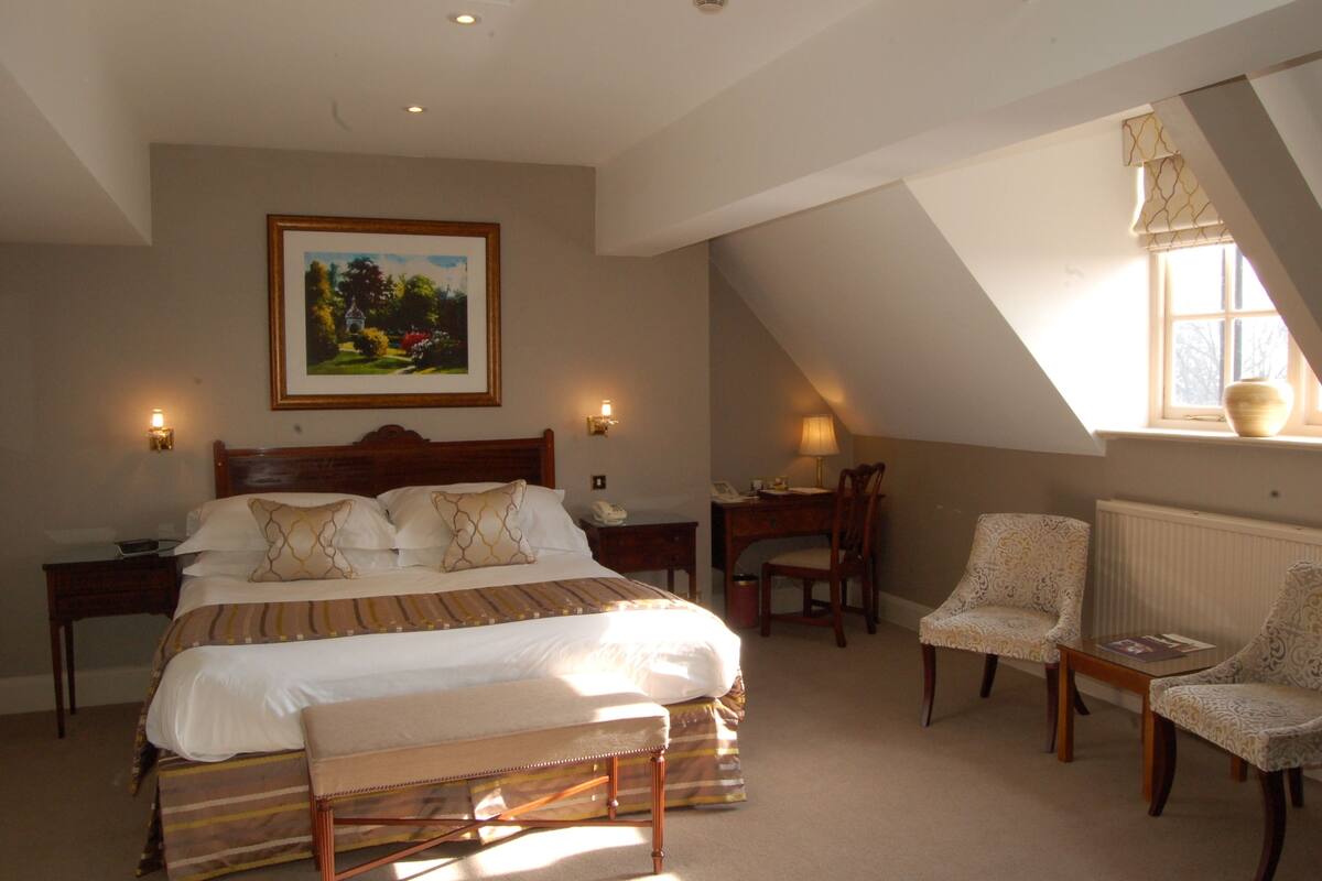 A bedroom with a large bed, two bedside tables, a desk with a chair, and a window with a view of trees.