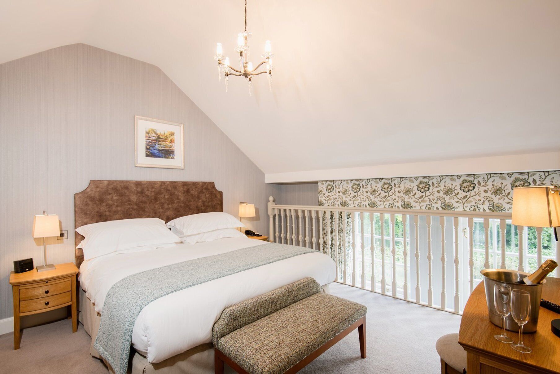 A bedroom with a large bed, a bench, a nightstand, a lamp, a chandelier, a framed picture, and a window with a view.