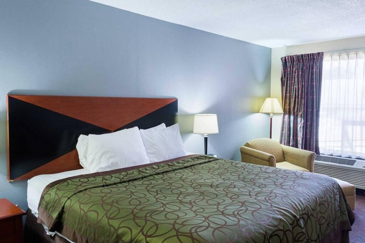 Une chambre d’hôtel avec un lit, une chaise, une lampe et une fenêtre avec des rideaux.